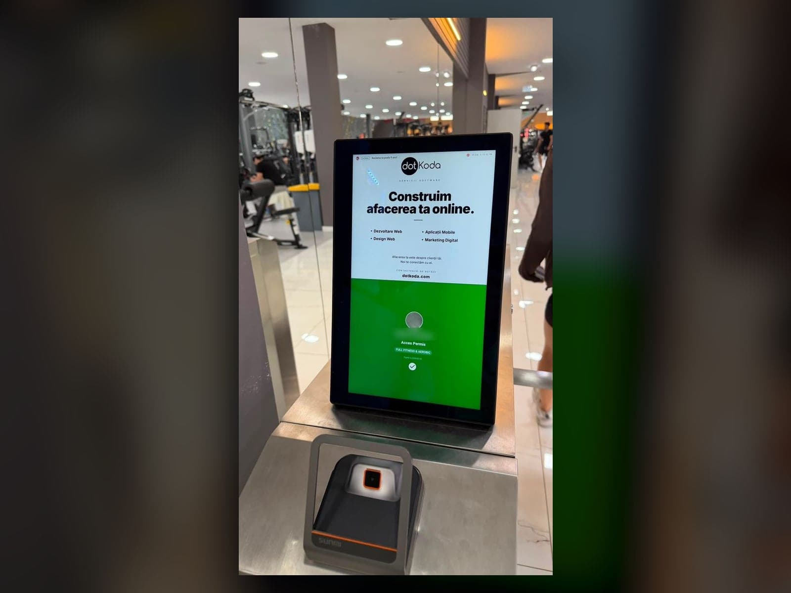 Turnichet XPE într-o sală de fitness: tableta afișează o reclamă în partea de sus și ecranul verde de acces permis dedesubt, cu scannerul de coduri QR montat pe carcasă